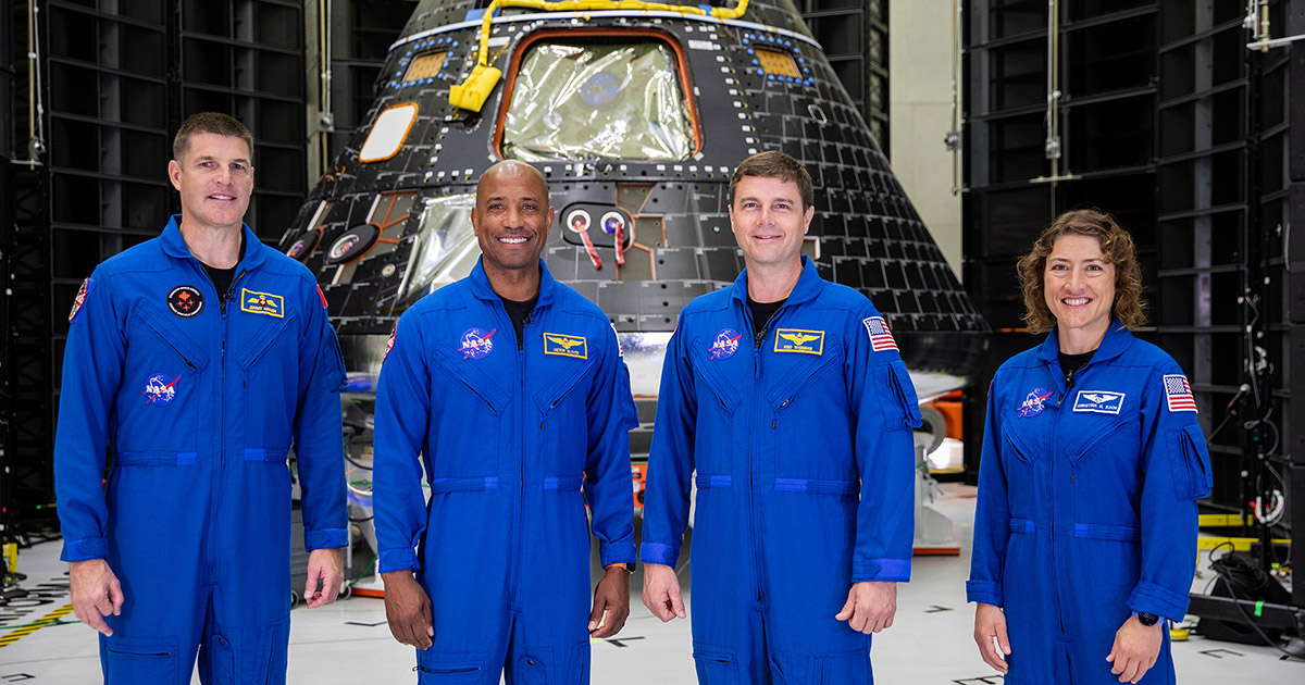 Four astronauts wearing blue flight suits pose in front of a tear-shaped capsule.