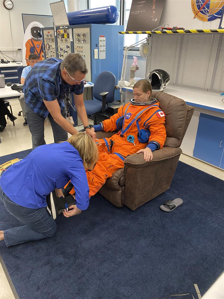 Jenni is sitting in an armchair while two people help her put on her spacesuit. 