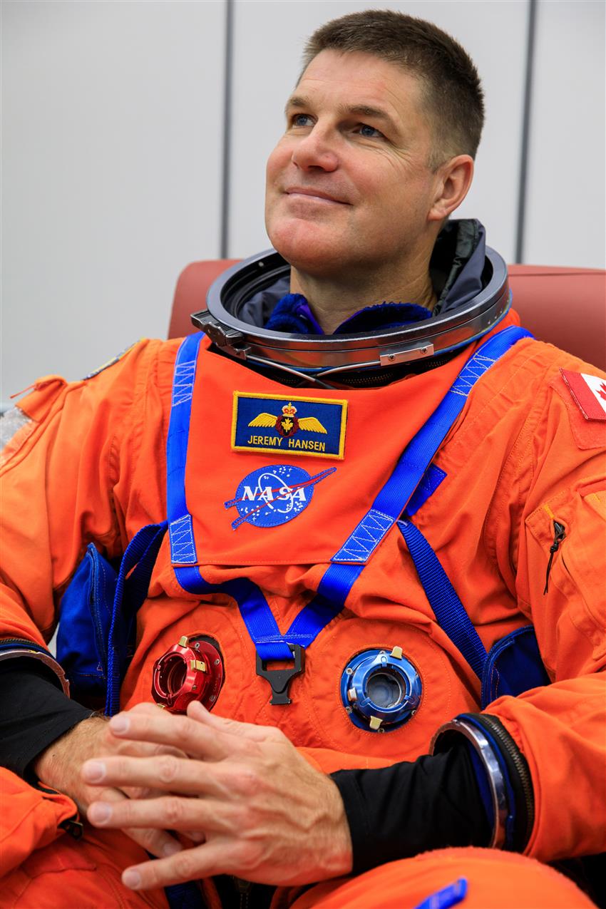 Astronaute souriant assis sur une chaise et vêtu d'une combinaison spatiale orange.