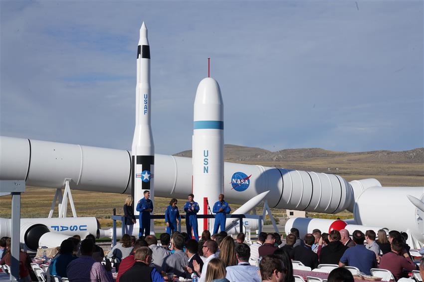 Quatre astronautes et une femme sont sur une estrade devant un public. À l’arrière-plan, il y a des fusées. 