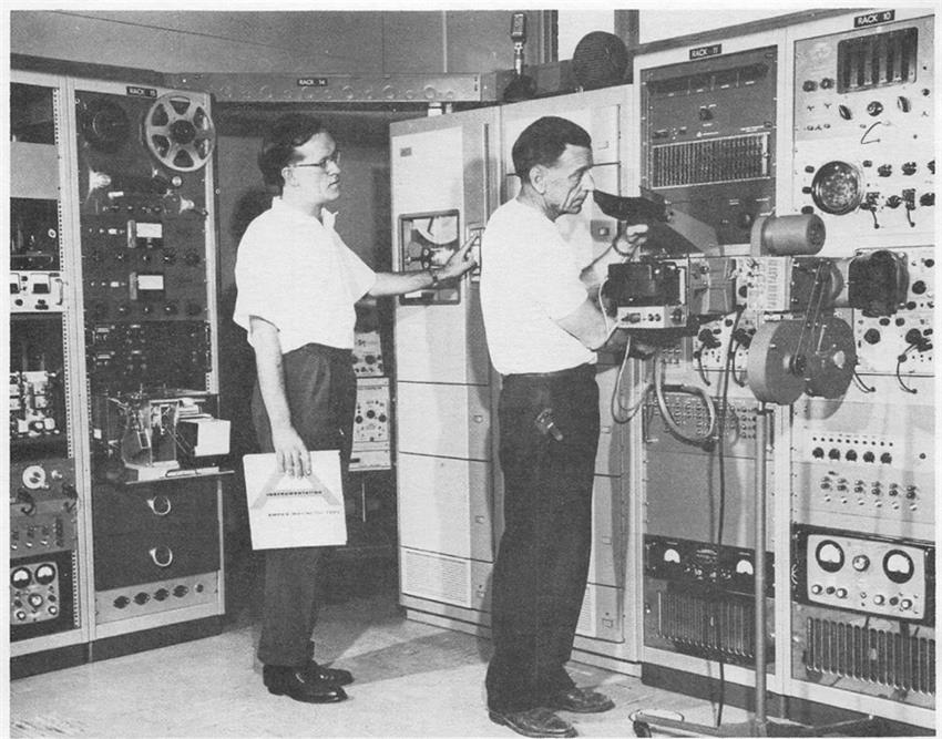Two men stand in front of electronic equipment.