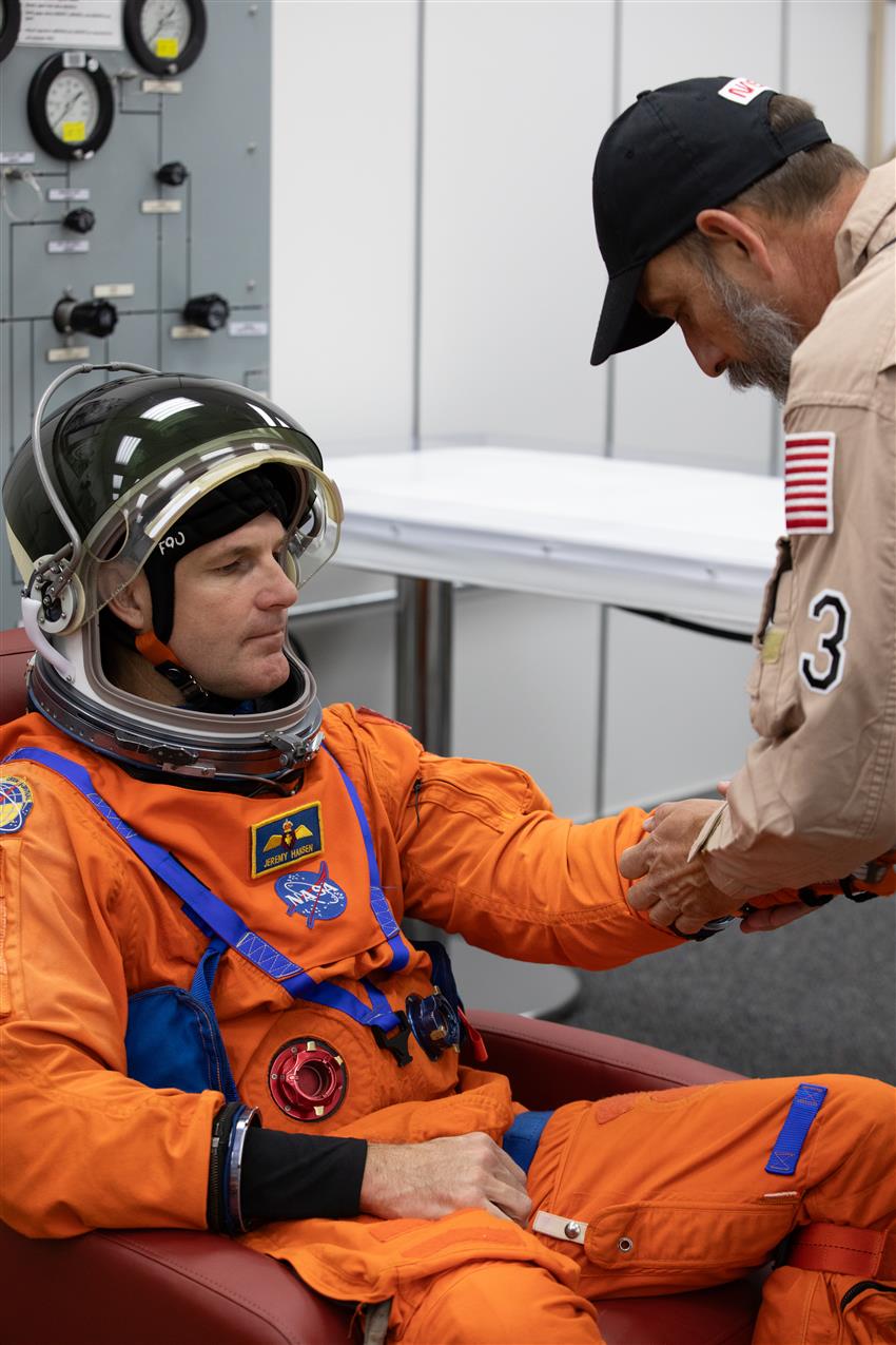An astronaut is wearing an orange spacesuit, and a man helps him adjust the sleeves.