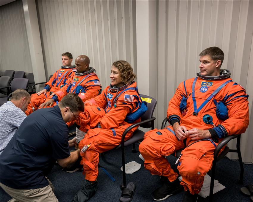 Quatre astronautes vêtus de leur combinaison spatiale sont assis, et deux personnes les aident à mettre leurs bottes.