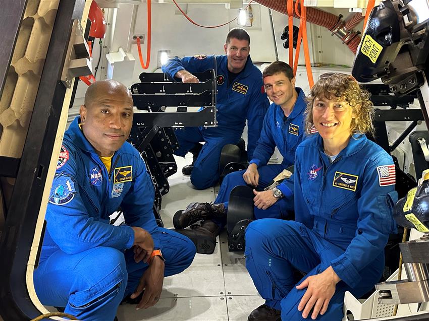 Four astronauts wearing blue flighsuits crouch down and pose inside a small spacecraft.