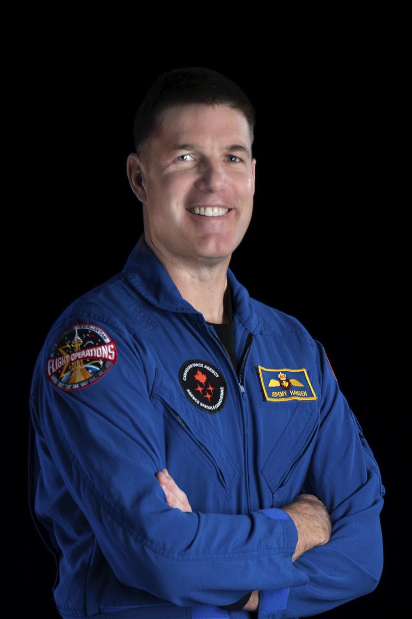 Jeremy Hansen stands facing the camera, he is wearing a blue flight suit