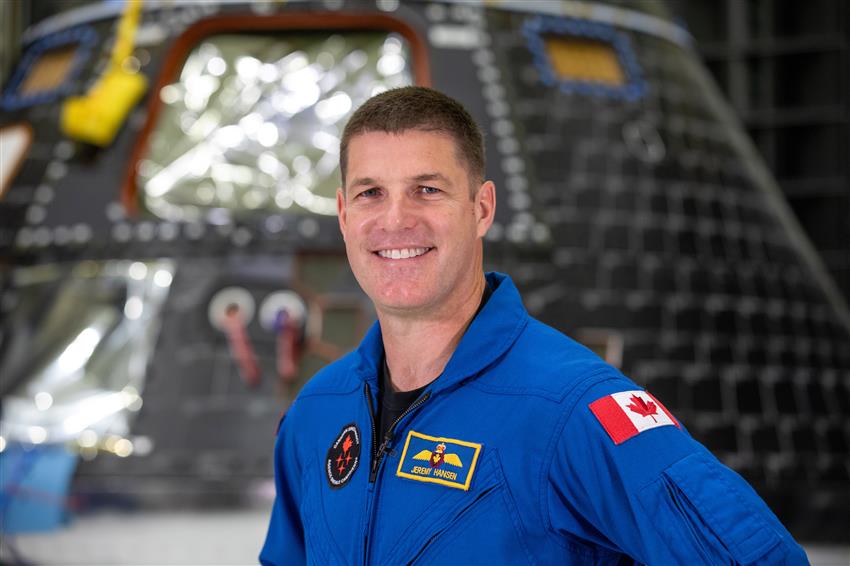 An astronaut wearing a blue flight suit smiles in front of a tear-shaped capsule.