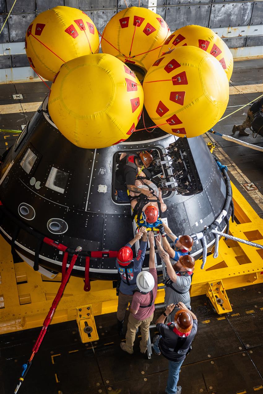 People loading a mannequin into a full-scale mock-up of a tear-shaped spacecraft.