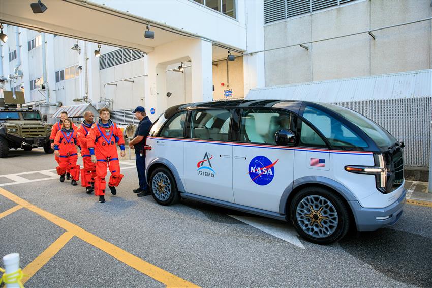 Four astronauts in orange spacesuits standing next to a van.