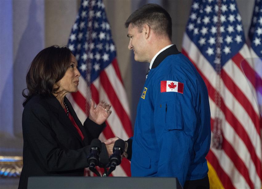 Jeremy Hansen wearing his flight jacket shakes hands with Kamala Harris. There are several American flags behind them.