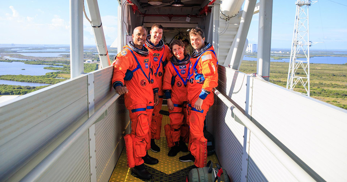 Four astronauts in orange spacesuits pose on a metal walkway high in the air