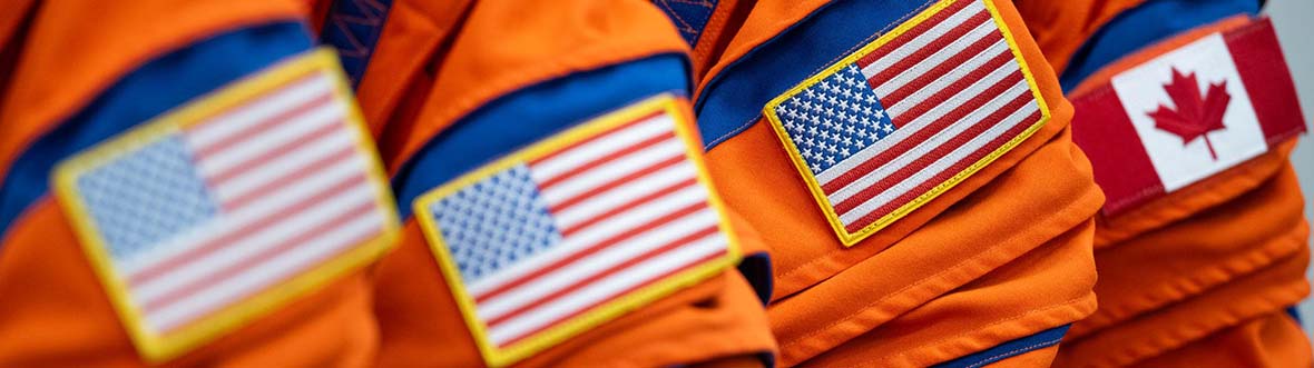 Close-up of spacesuits hanging on a rack, three with an American flag and one with a Canadian flag.