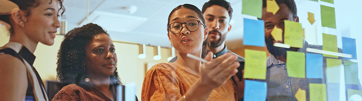 Woman standing in front of glass wall with sticky notes, talking to teammates listening attentively