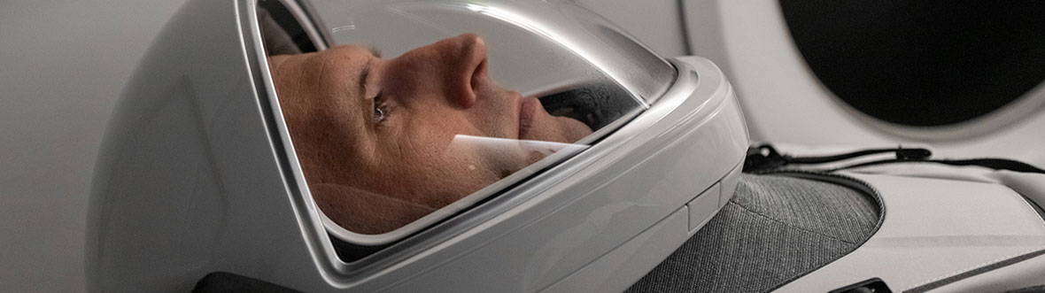 Joshua Kutryk is lying on his back wearing a spacesuit and helmet in a spacecraft simulator.