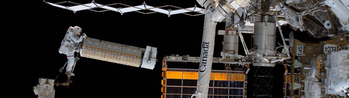 Sur fond d'obscurité spatiale, un astronaute en combinaison déplace un panneau solaire vers la Station spatiale.