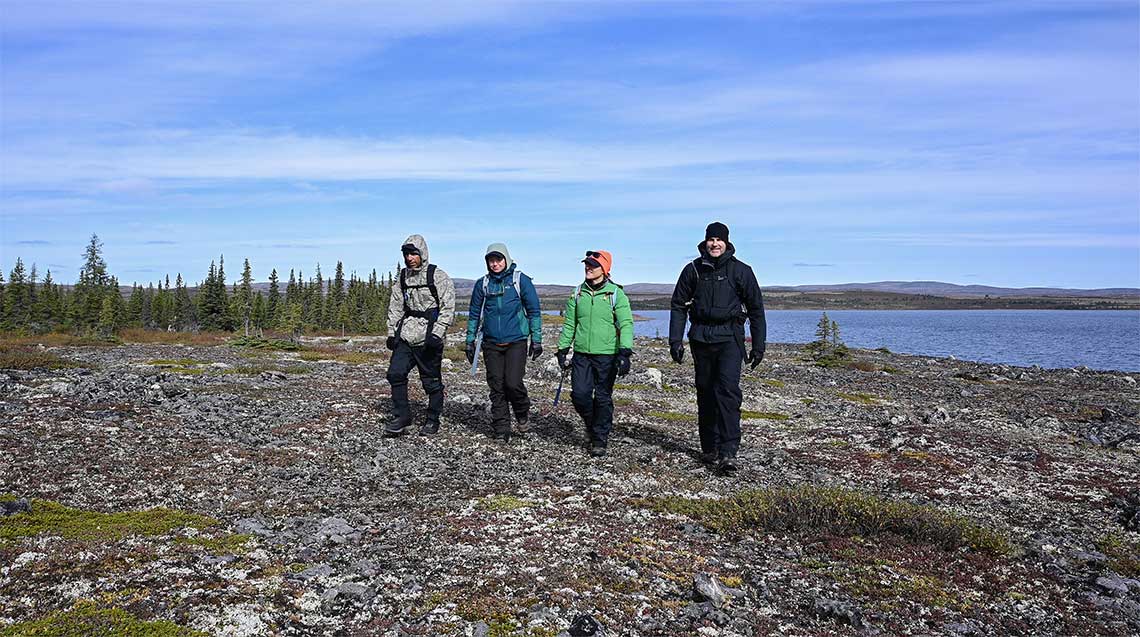 Four people in warm gear walk on rocky terrain surrounded by tundra and a lake behind them.