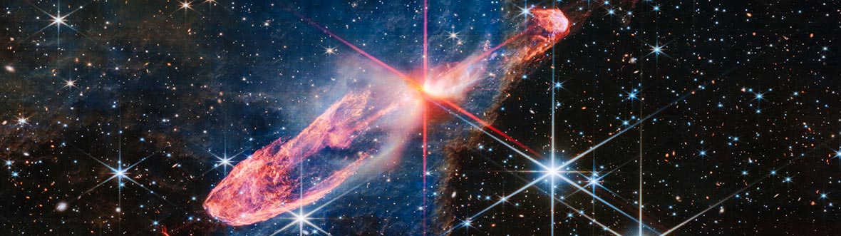 Colorful cosmic scene showing a bright young star surrounded by glowing jets of pink, red, and blue gas set against a dense field of stars in deep space.