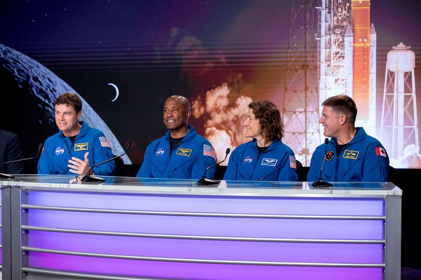 Four astronauts wearing blue flight suits sit side by side behind a table with microphones.