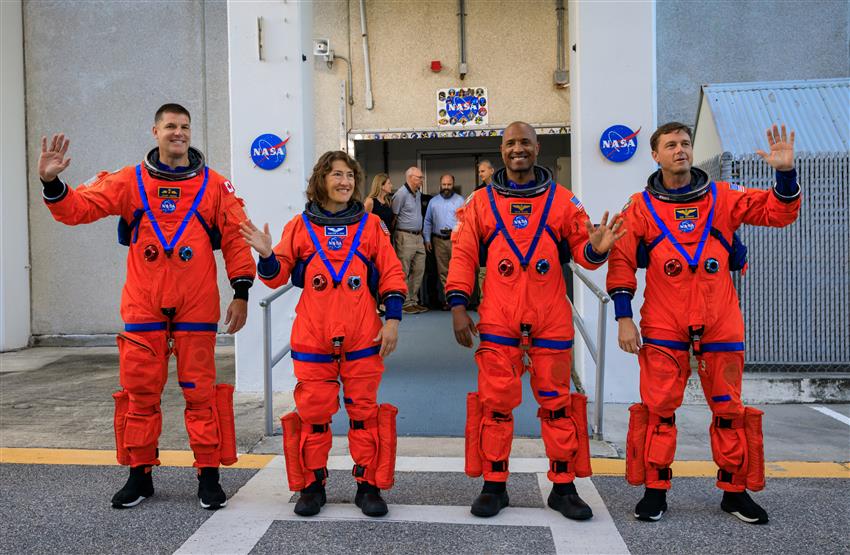 Quatre astronautes vêtus de combinaisons spatiales orange font un signe de la main devant un bâtiment.