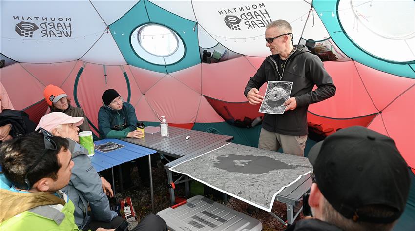 A man shows an image of a crater to five people sitting around a table. They are in a tent.