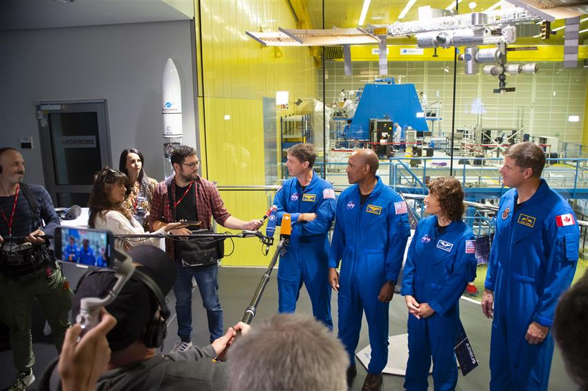 Four astronauts in blue flight suits being interviewed and filmed by a group of journalists.