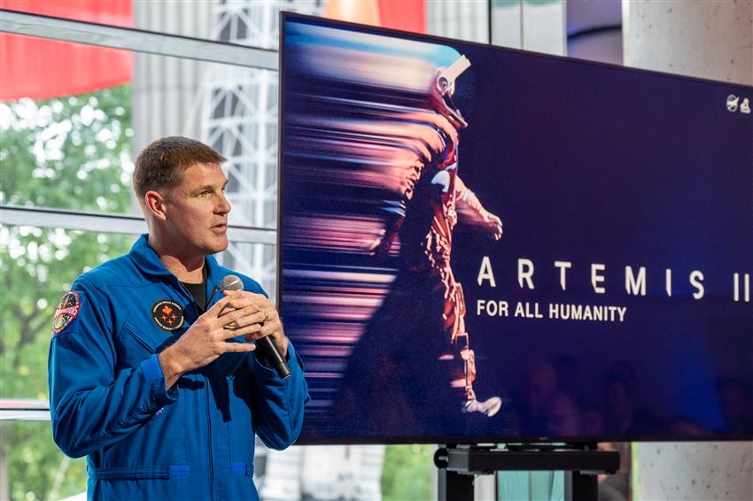 Jeremy Hansen a un micro à la main, derrière lui, sur un écran est projetée une infographie d'un astronaute