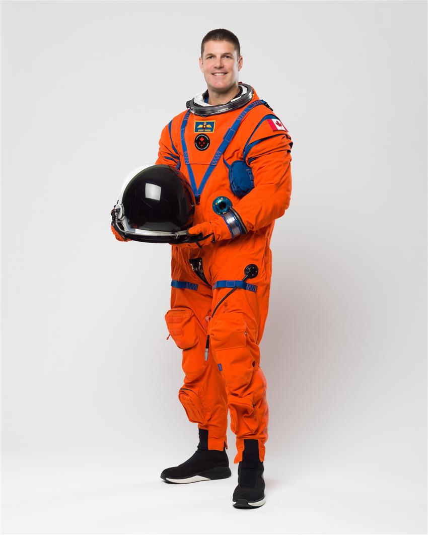 Jeremy Hansen stands in front of a white wall in his orange Artemis II flight suit
