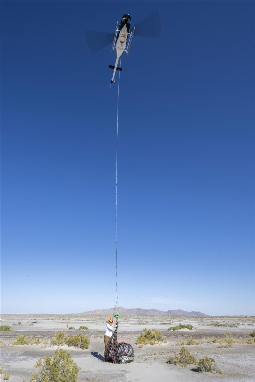 A person is attaching a package containing a space capsule to a wire hanging from a flying helicopter.