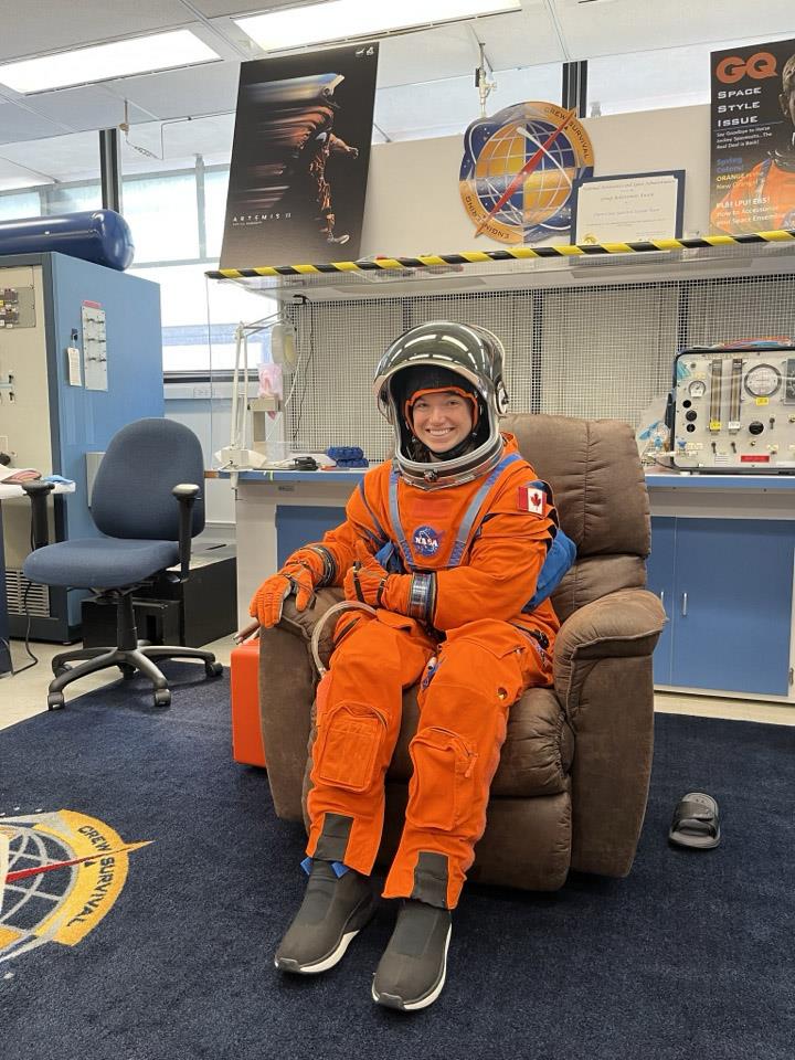 Jenni is wearing an orange spacesuit, sitting in an armchair and smiling.