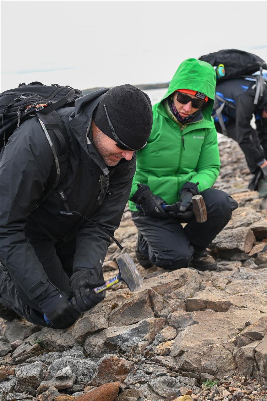 Two people in warm clothing kneel with rock hammers in their hands. One of them is hitting the rocky ground.