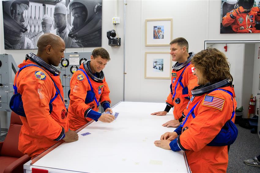 Four astronauts stand around a table. One of them is distributing playing cards.