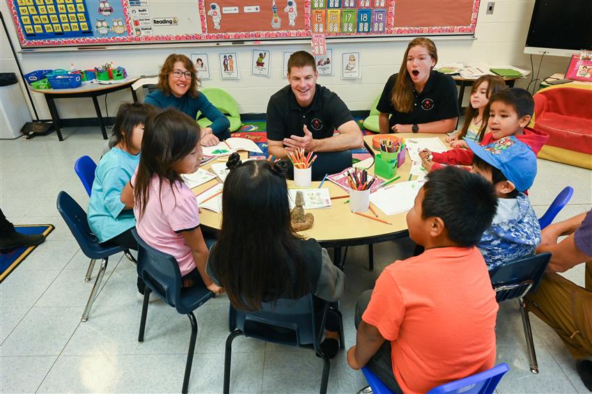 Trois adultes et sept enfants sont assis autour d’une table, dans une salle de classe.