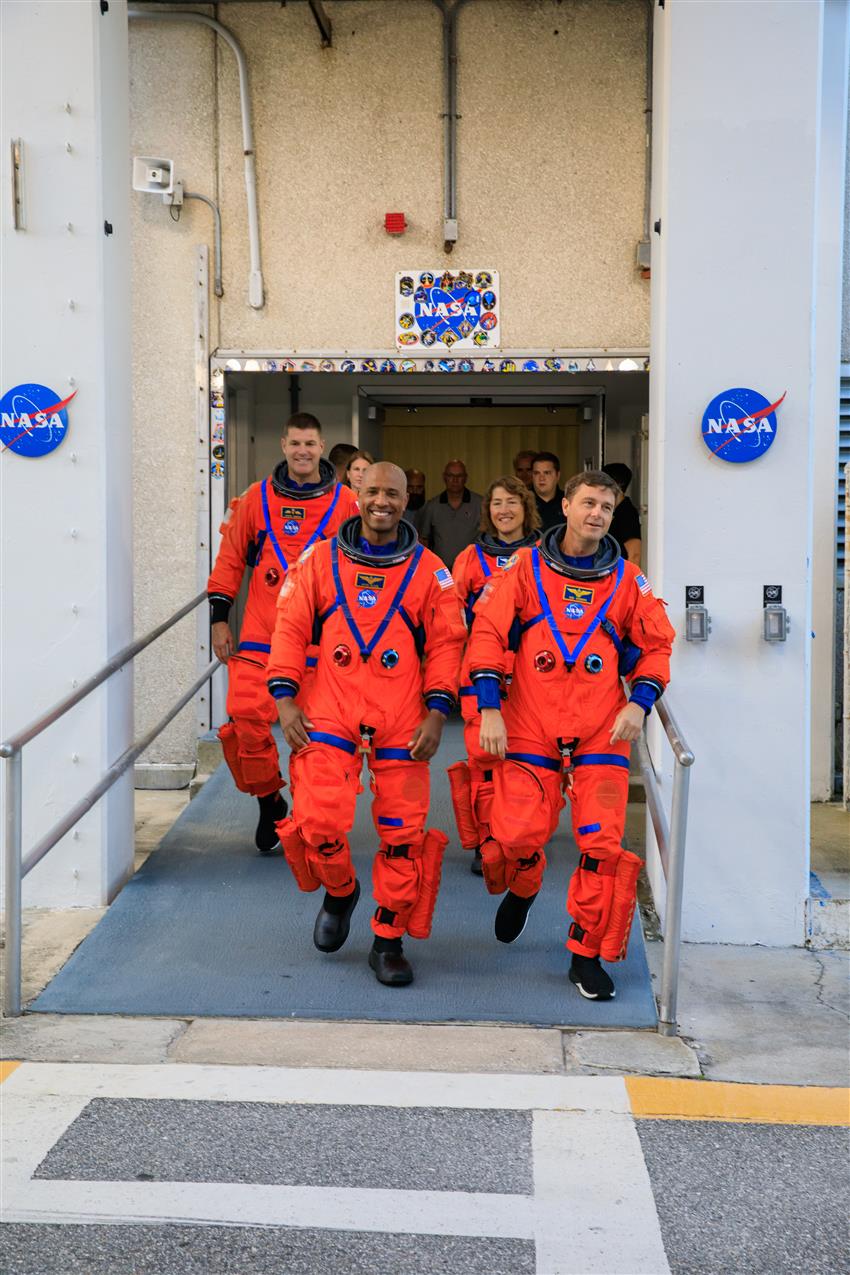 Four astronauts in their orange spacesuits step out of a building.