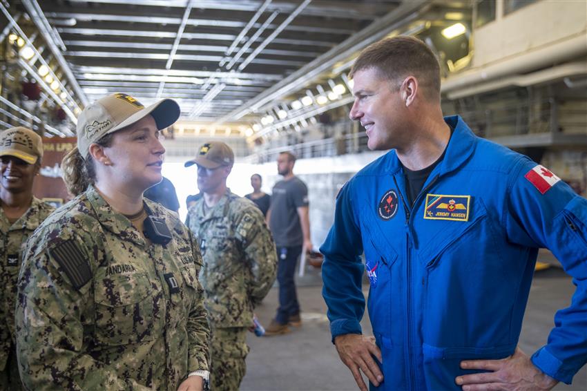 Une membre de la Marine américaine en uniforme discute avec Jeremy Hansen qui sourit et porte une combinaison bleue.