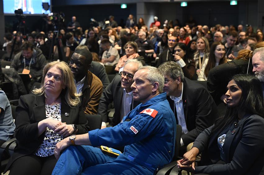 Une foule de gens regarde un écran. Sherry, David et Kumi sont assis au premier rang.