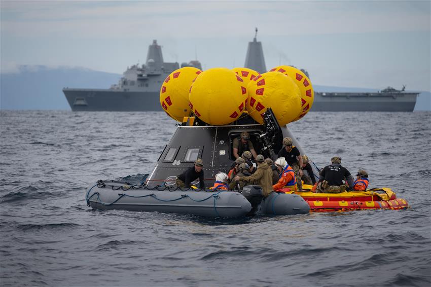 Two inflatable boats carrying people float in front of a cone-size mockup of a spacecraft. 