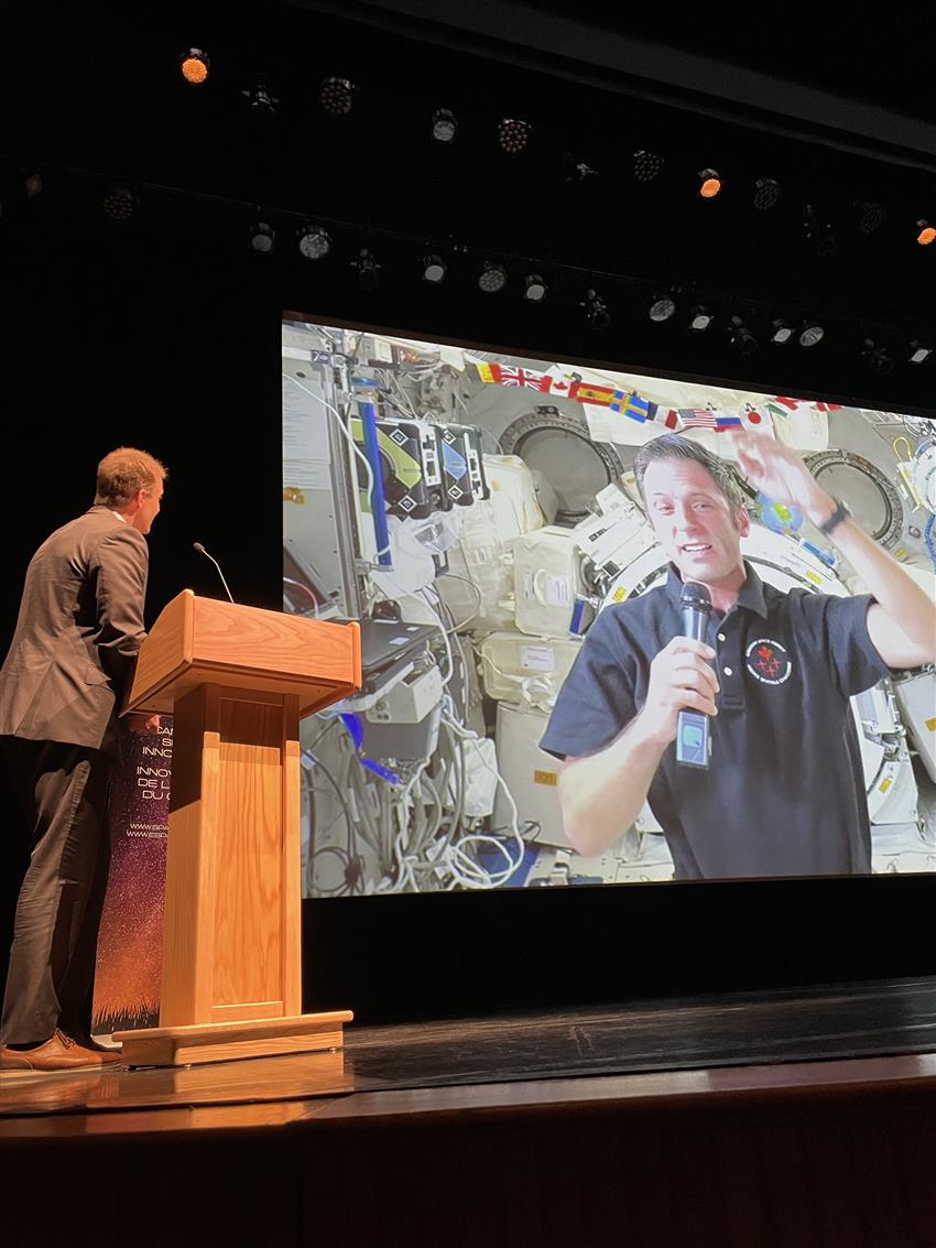 Joshua appears on screen, speaking live from the International Space Station. Brian is standing next to the screen.