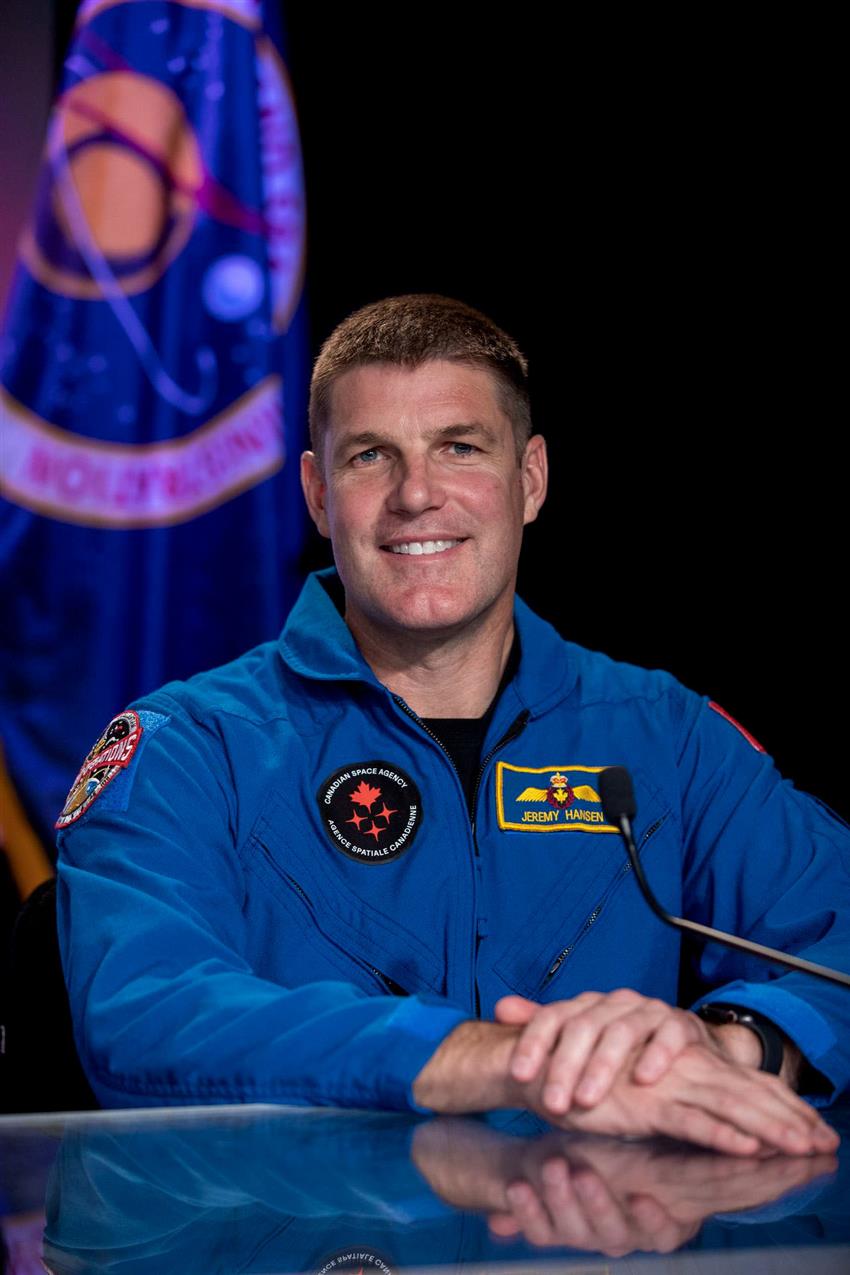 An astronaut dressed in his blue flight suit sits at a table.