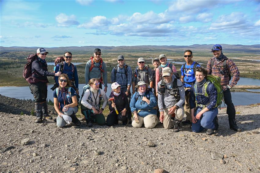 Fifteen people over two rows pose for a group photo with a lake and the tundra in the background.