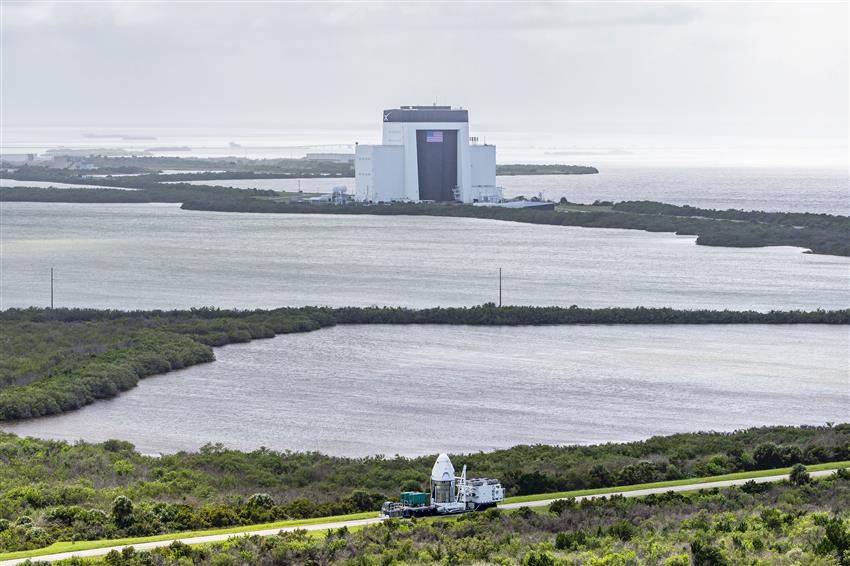 A large hanger, surrounded by water, and the Dragon vehicle on a transport vehicle is seen on a road. 