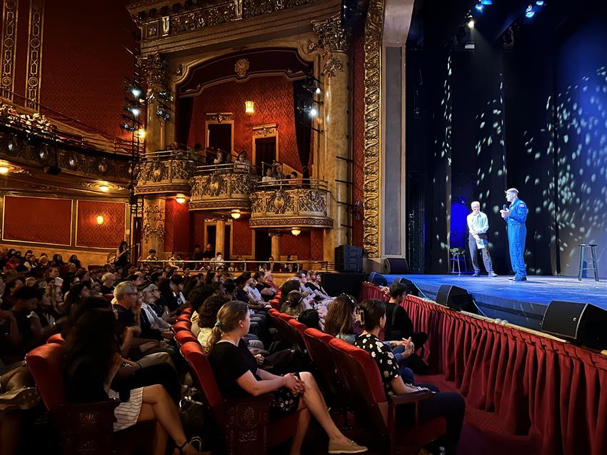 Jeremy and Paul stand on stage in an old, elegant theatre full of people.