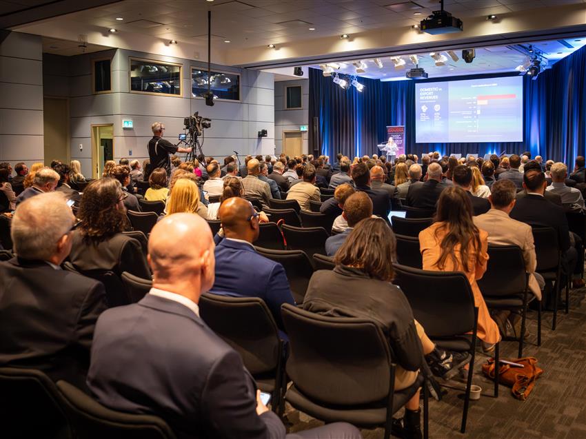 Plusieurs personnes dans une grande salle écoutent une conférencière. Derrière elle, un écran géant.