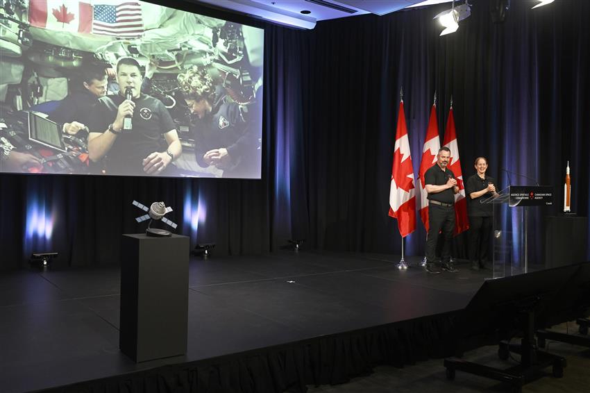 Jeremy and the other crewmembers appear on a big screen. Jamie and Tim stand at a podium.