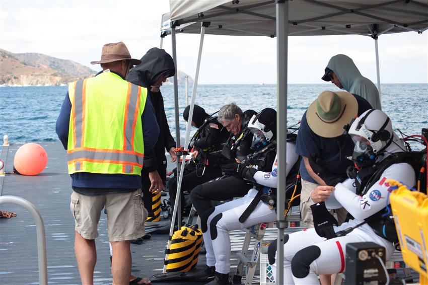 Josh and Marcos, in diving suits, check their equipment while others assist. 