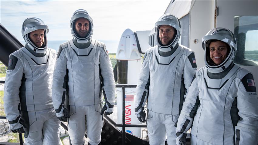 The four astronauts stand in a line in their space suits. A rocket is visible behind them. 