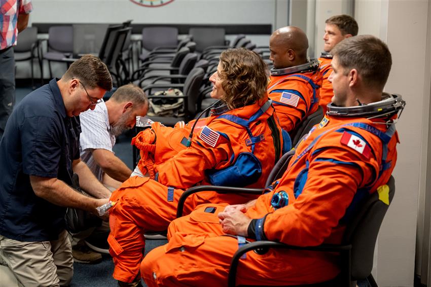 Four astronauts are wearing orange spacesuits, and two people help them put on their boots.