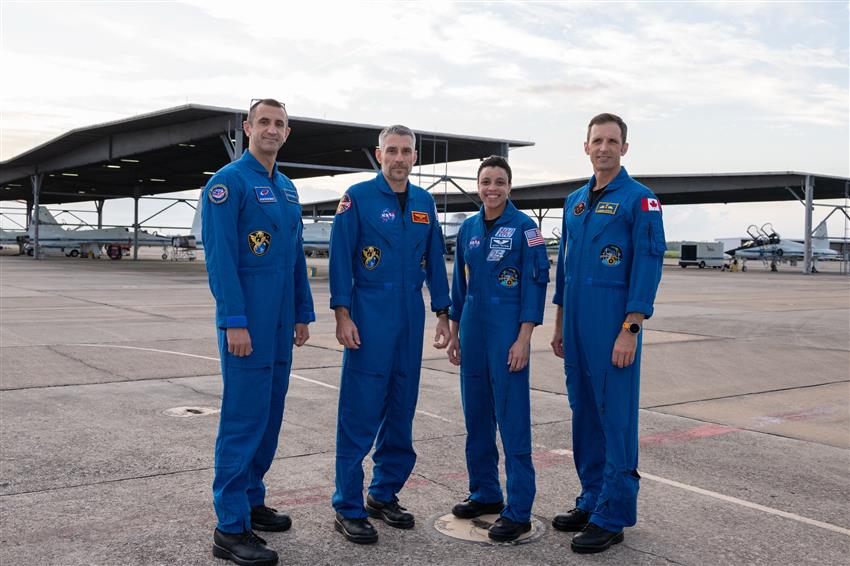 Sergey, Luke, Jessica, and Josh stand in a line with airplanes behind them. 
