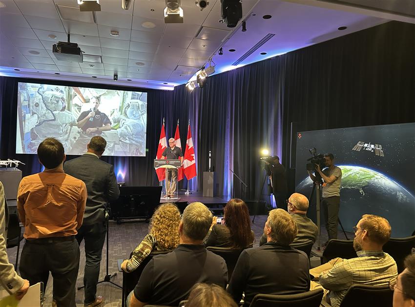Joshua appears on screen, speaking live from the Space Station, while journalists and guests attend the event.