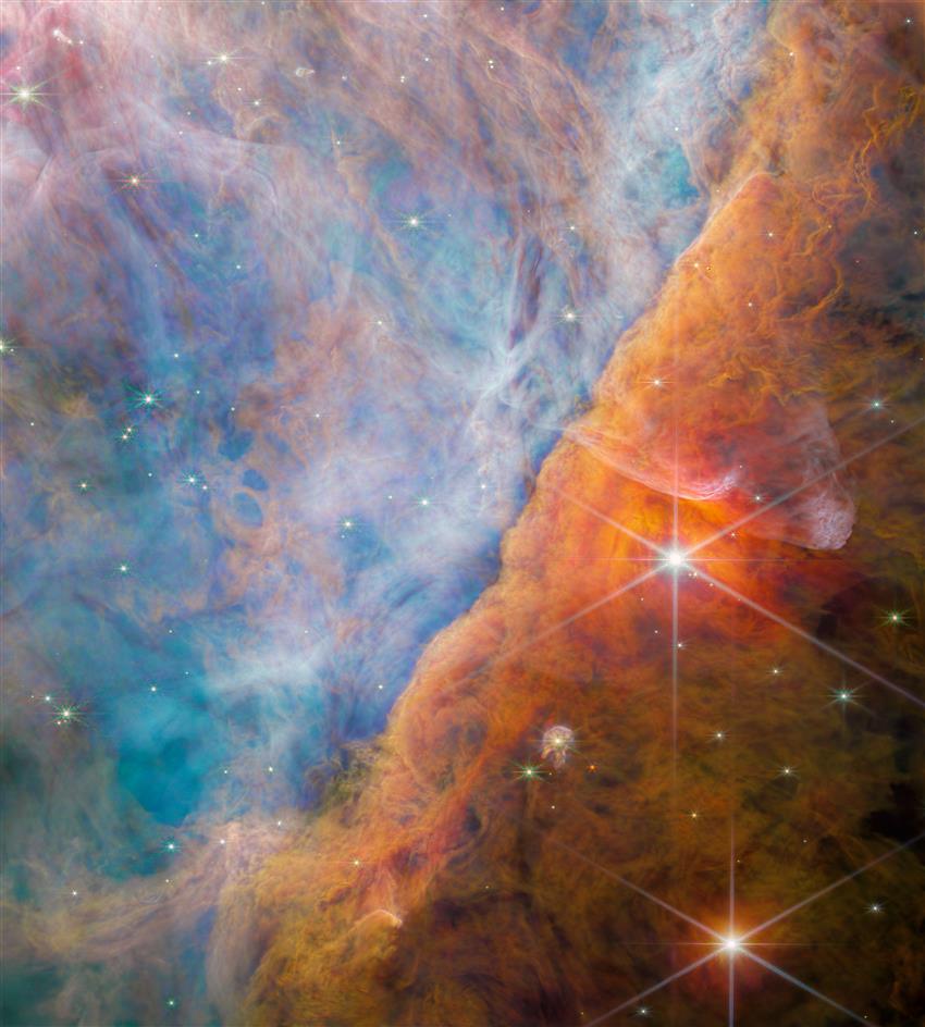 Colourful image of part the Orion nebula, with numerous stars shining brightly through the dust and gas.