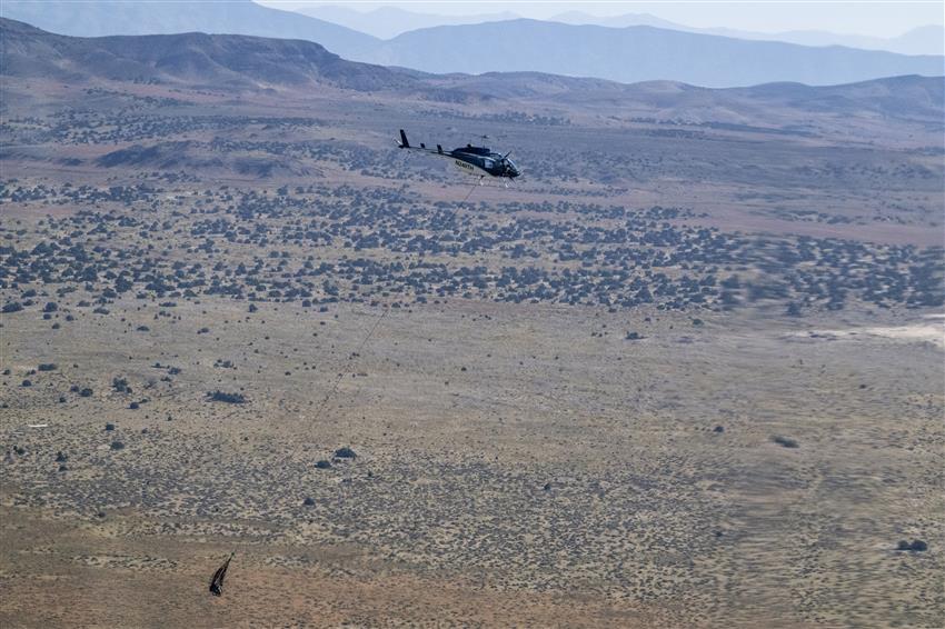 A helicopter transports a space capsule over the desert.