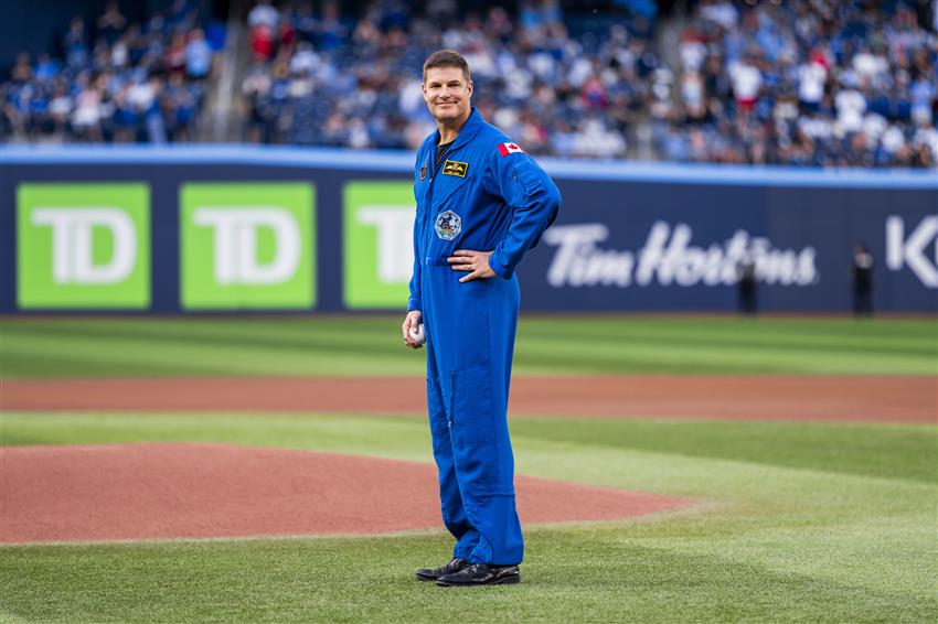 Jeremy is standing on a field with a baseball in his right hand. He is wearing his flight suit. 